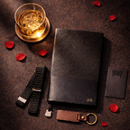 Personalised Black leather journal, keychain, bottle opener, glass of whiskey, sleeve garter, and rose petals on a dark surface