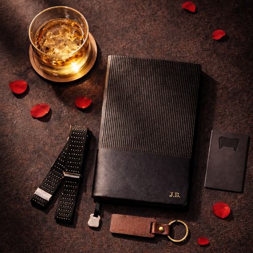 Personalised Black leather journal, keychain, bottle opener, glass of whiskey, sleeve garter, and rose petals on a dark surface