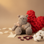 Plush teddy bear with a heart-shaped tag next to a red knitted heart on a beige background.