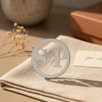 Silver monogrammed decorative piece on a beige fabric surface with a blurred background